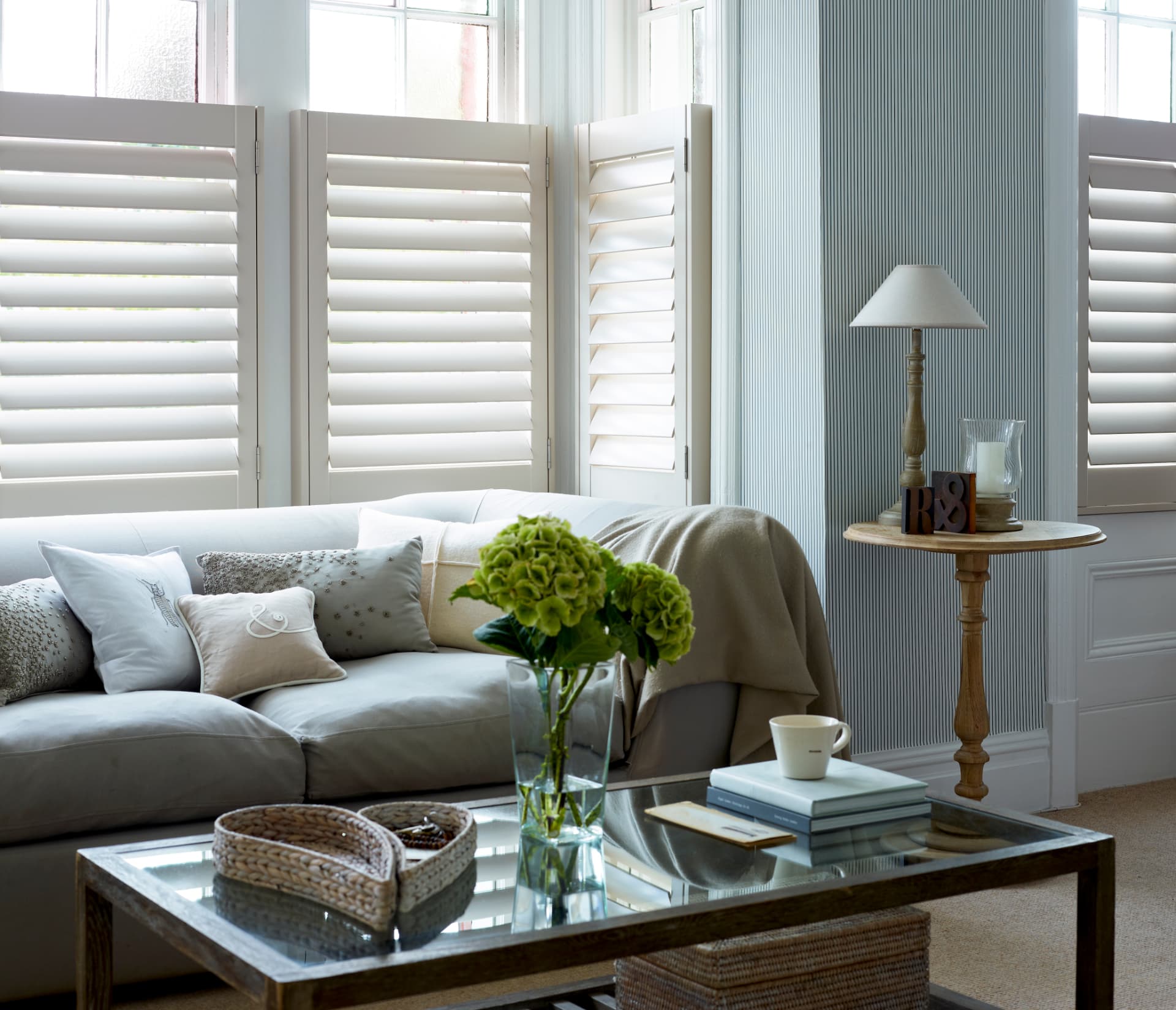 Soft white café style shutters in a bay window above a linen sofa